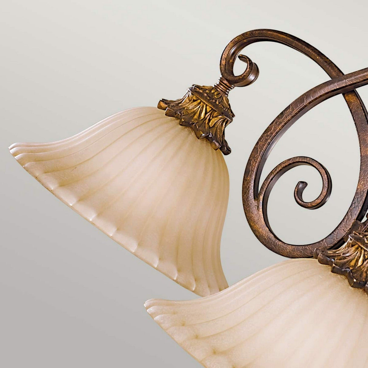 Close-up view of the Sonoma Valley 3 Light Chandelier showcasing rustic elegance with two bell-shaped, frosted glass shades. The metalwork features intricate scroll and leaf designs in a rich bronze finish, reminiscent of French Country antiques, all set against a neutral background.