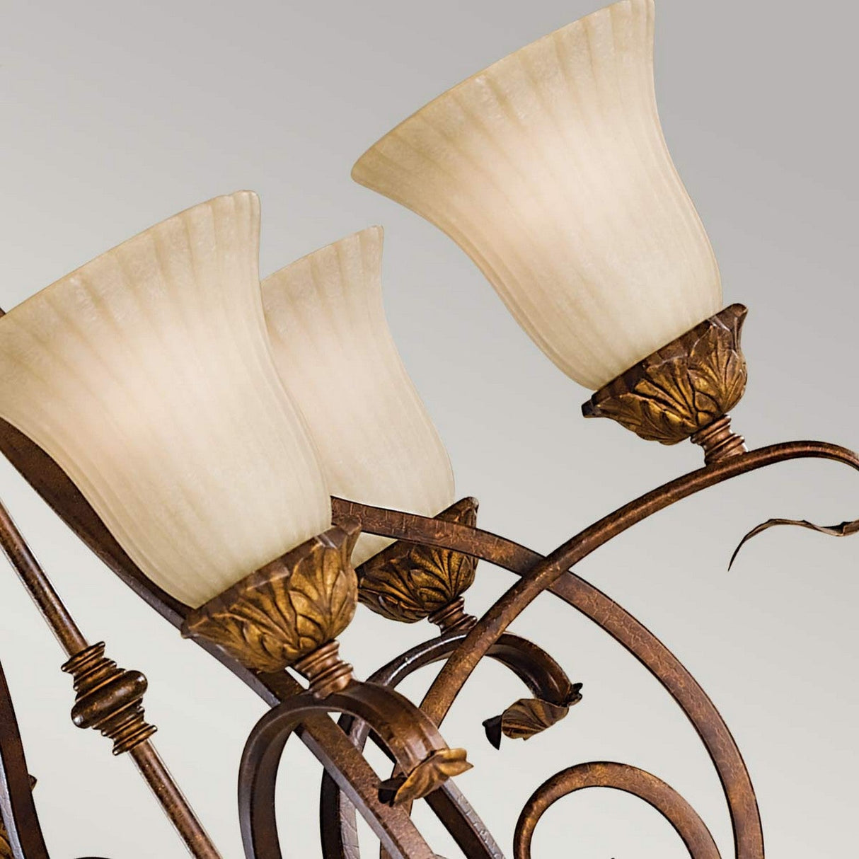 A close-up of the Sonoma Valley 6 Light Chandelier in cream and bronze highlights its opal glass shades, ornate bronze metalwork adorned with leaf motifs, against a soft gray background. Its design resembles French Country antiques with three fluted, cream-colored lampshades.
