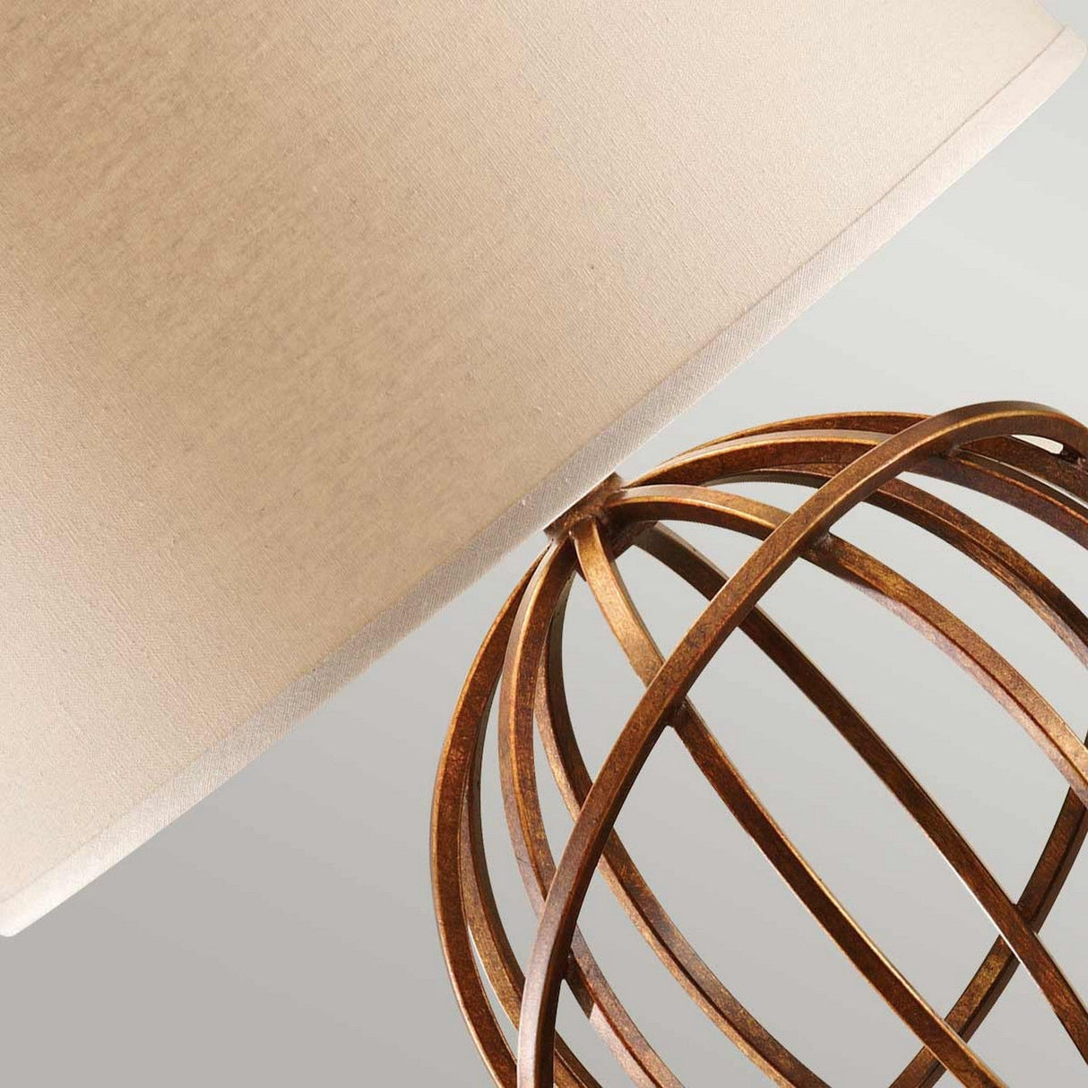Close-up of the Spencer 1 Light Table Lamp in Firenze Gold, featuring a spherical metal globe structure paired with a beige fabric shade. The design highlights a rust-colored finish on the metal, captured from an angled perspective against a neutral backdrop.