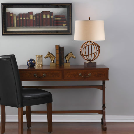 A wooden desk with two drawers is adorned with horse head bookends, stacked books, a decorative globe featuring a metal structure, and a sleek Spencer 1 Light Table Lamp in Firenze Gold. Above hangs a framed painting of books. A black leather chair sits in front on the dark walnut base floor.