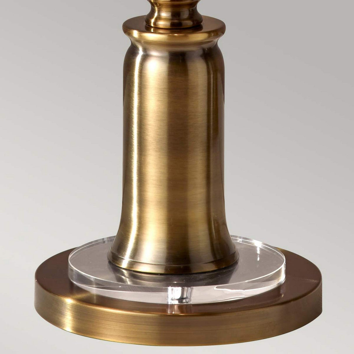 The Stateroom 2 Light Floor Lamp - Brass showcases a close-up of its brass cylindrical base with a clear acrylic ring at the midpoint. This sophisticated lamp is anchored by a circular platform, both of which reflect light with a gentle, metallic glow against a monochromatic backdrop.