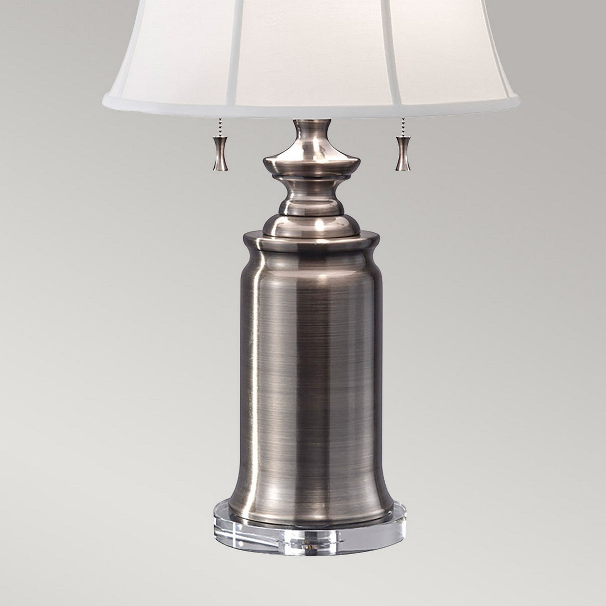 The Stateroom 2 Light Table Lamp in Antique Nickel, featuring a brushed metal finish with a cylindrical base and a cotton linen shade, offers versatile lighting through convenient pull chains, making it an elegant addition against any neutral backdrop.