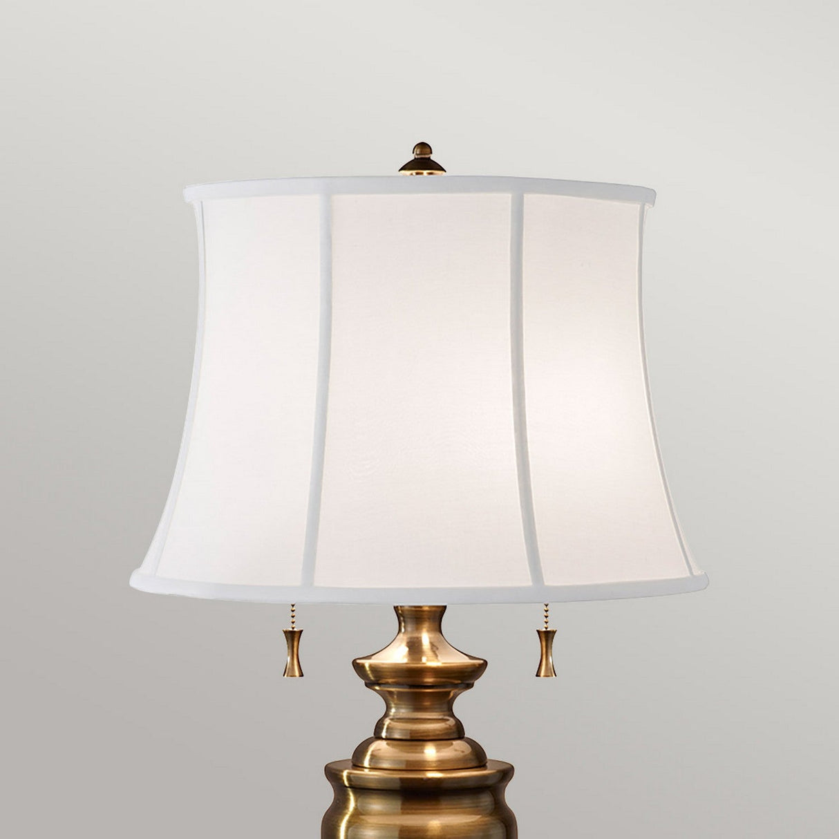 The Stateroom 2 Light Table Lamp - Brass showcases a white, rectangular lampshade set against a simple gray background. Featuring two pull chains and an ornamental finial atop, it's an elegant touch to enhance any home decor lighting arrangement.