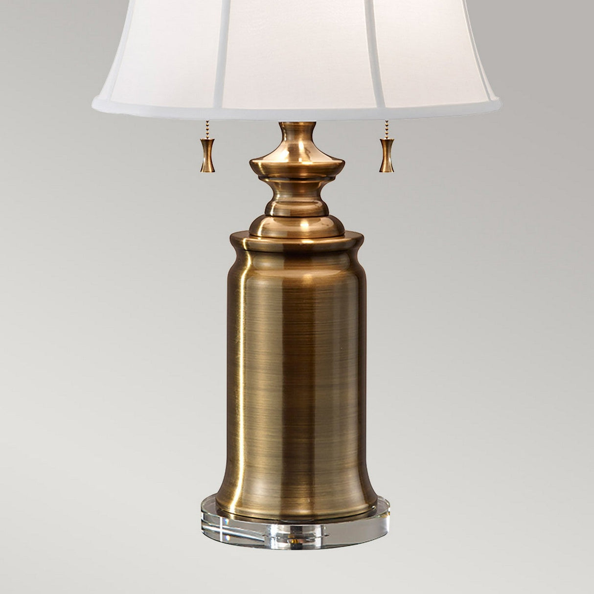 The Stateroom 2 Light Table Lamp - Brass features a round clear base and a white fabric lampshade. Its classic design is enhanced by decorative pull chains beneath the shade, making it an ideal addition to any home decor lighting collection.