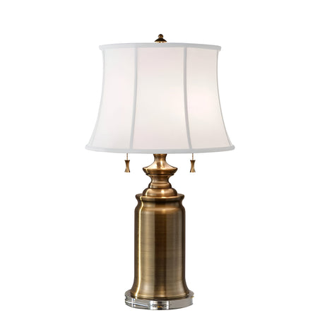 The Stateroom 2 Light Table Lamp - Brass features a tall, cylindrical base paired with a wide, white fabric lampshade. This sophisticated home decor piece includes two pull chains adorned with small tassels and is accented with a decorative finial, combining functionality with style.