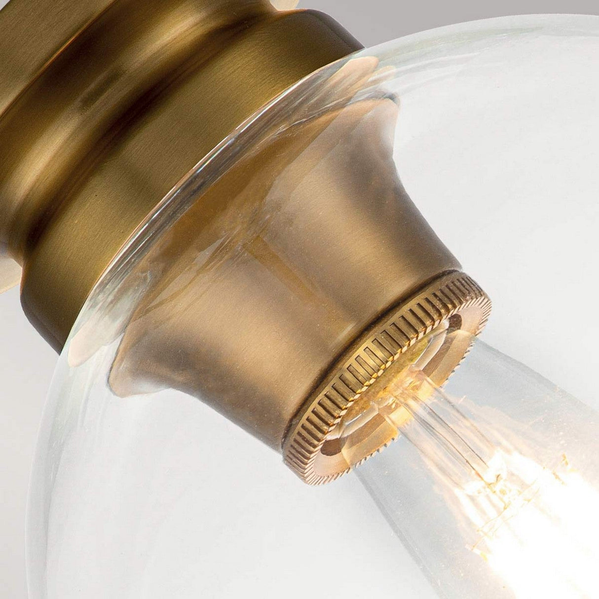A close-up view of the Tabby 1 Light Flush in Burnished Brass showcases a vintage-style light bulb encased in a clear glass fixture. The illuminated bulb casts a warm glow, highlighting the antique finish of the metal socket. This design elegantly features an exposed lamping aesthetic for an exquisite touch.