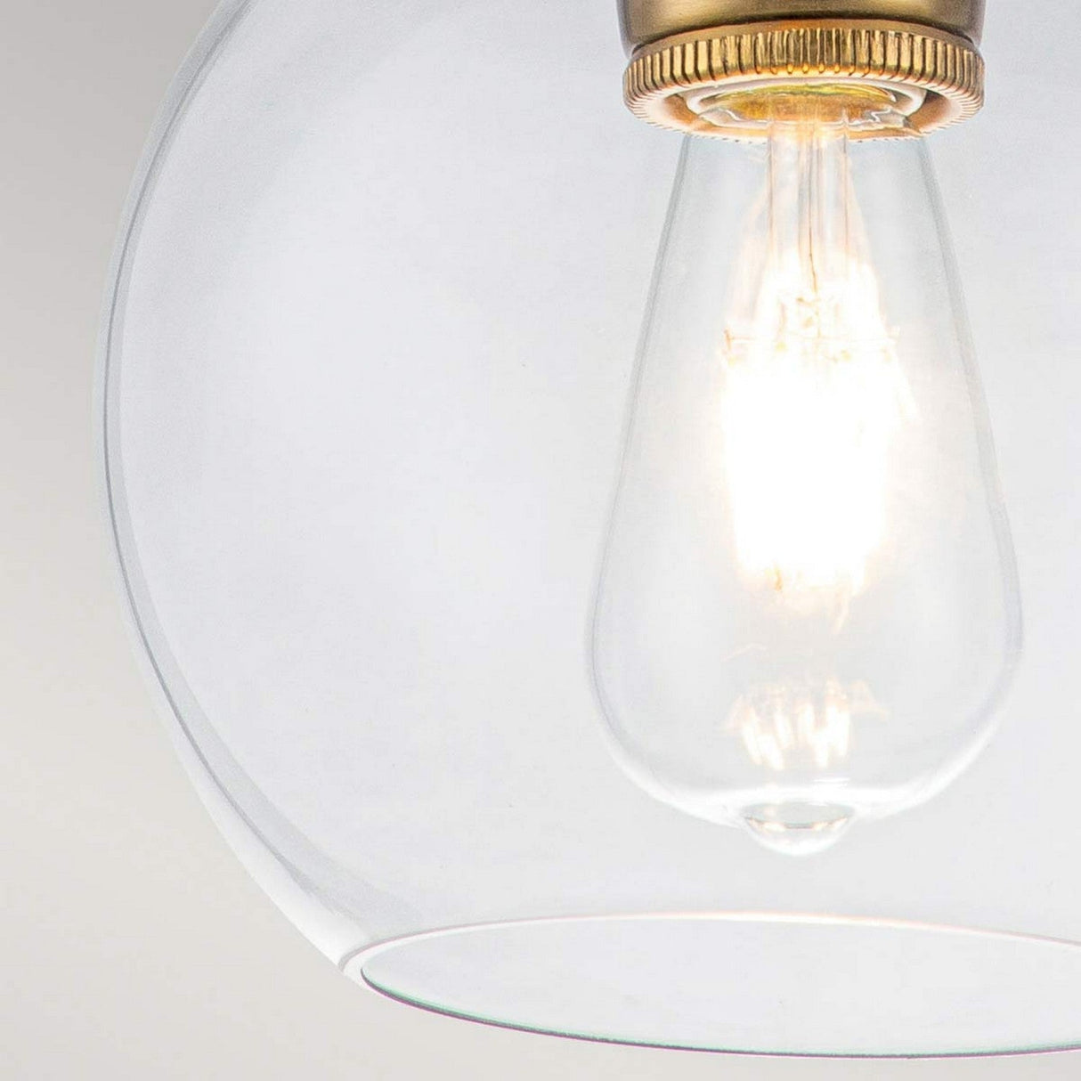 A close-up of the Tabby 1 Light Mini Pendant in a glass fixture highlights its lit filament, emitting a warm glow. The minimalist design is complemented by burnished brass accents, enhancing its classic elegance.