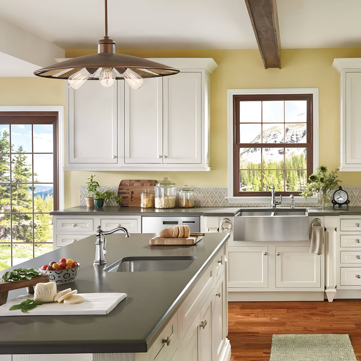 Bright kitchen with white cabinets, yellow walls, and large windows showcasing a scenic view of mountains and trees. The space includes a spacious island with a dark countertop, a farmhouse sink, and the Urban Renewal 3 Light Large Pendant in Bronze for an elegant yet modern touch.