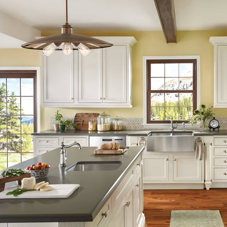 Bright kitchen with white cabinets, yellow walls, and large windows showcasing a scenic view of mountains and trees. The space includes a spacious island with a dark countertop, a farmhouse sink, and the Urban Renewal 3 Light Large Pendant in Bronze for an elegant yet modern touch.