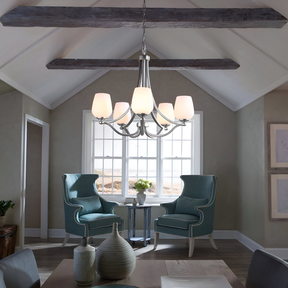 A cozy room with a vaulted ceiling featuring exposed wooden beams. Two green wingback chairs and a small table with flowers sit by a window. The Vintner 5 Light Chandelier - Satin Nickel, with its opal etched glass shades, illuminates the space adorned with vases and framed art.