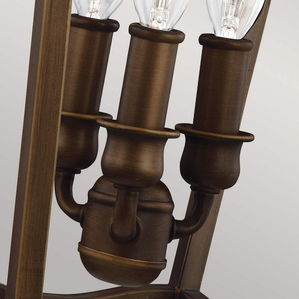 Close-up view of the Yarmouth 3 Light Chandelier, featuring a painted aged brass finish and three decorative candle-style bulb holders. The design is reminiscent of traditional lantern styles, with visible bulbs surrounded by the elegant bronze tone. The background is a plain, light gray.