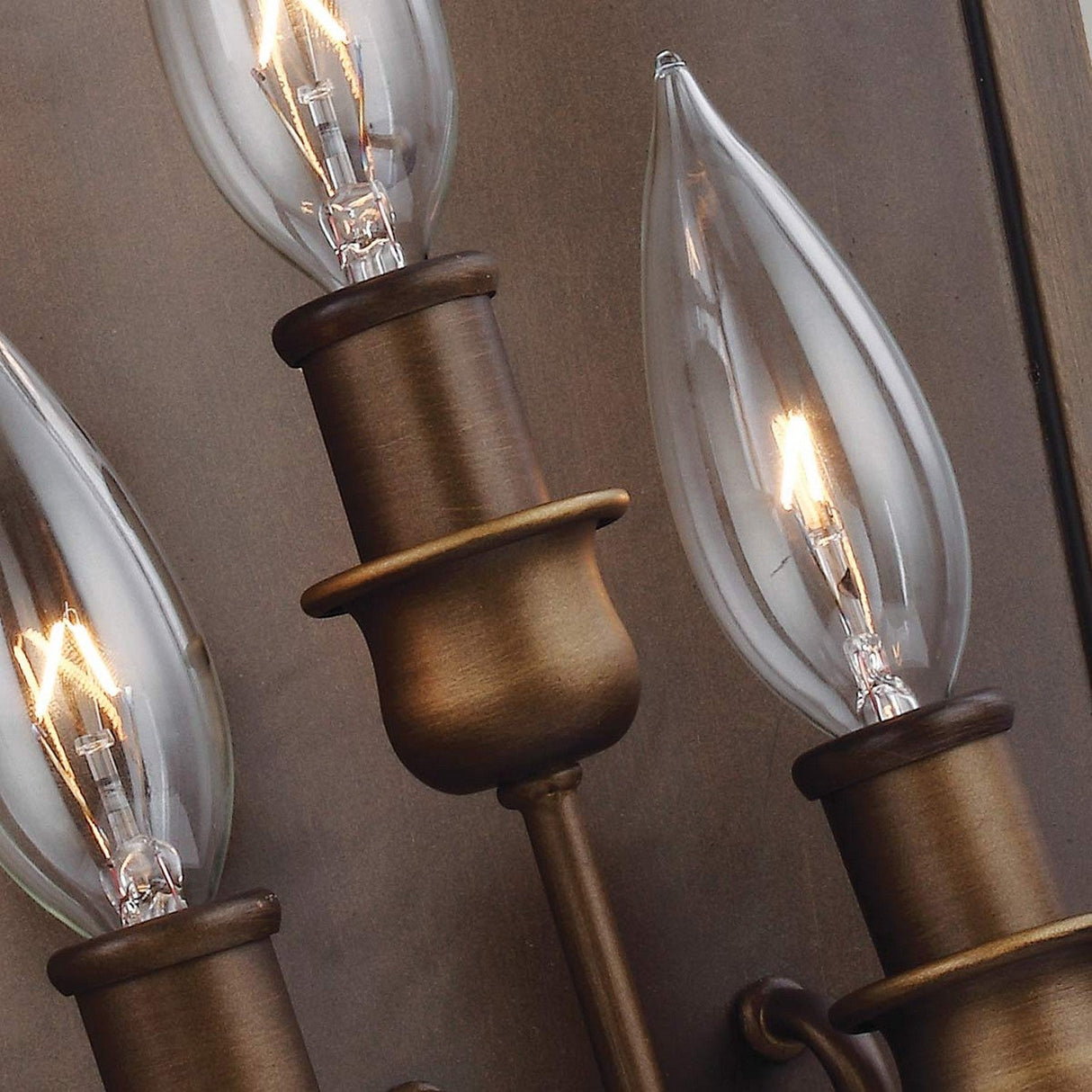 Close-up of vintage-style light bulbs with pointed glass tops glowing warmly, set in aged brass cylindrical holders. These are part of the Yarmouth 3 Light Wall Light - Painted Aged Brass fixture, evoking a design reminiscent of traditional lanterns.