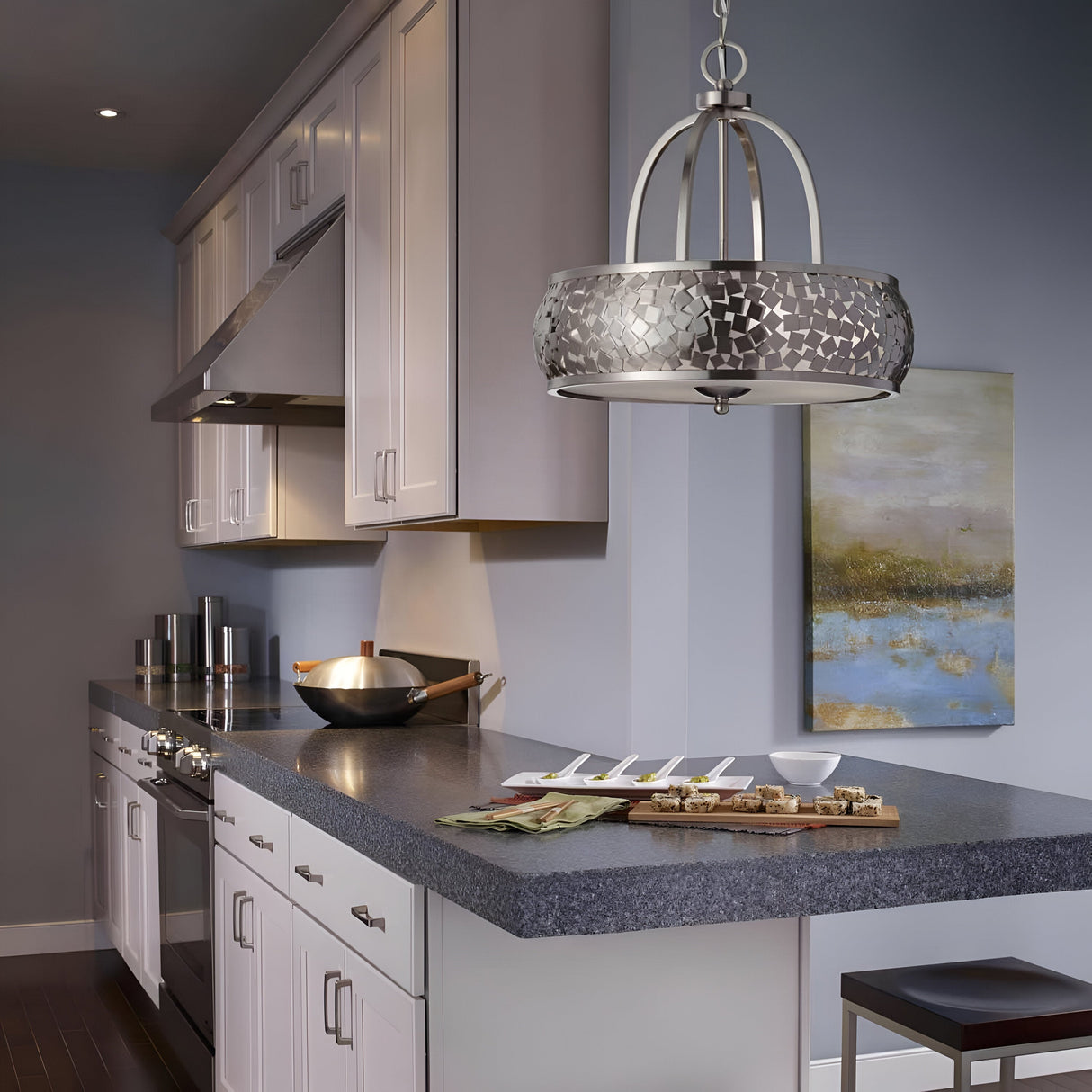 The modern kitchen features white cabinets, stainless steel appliances with a brushed steel finish, and a gray countertop. Above the counter with appetizers and a wok hangs the Zara 4 Light Chandelier in brushed steel. A landscape painting on the light blue wall enhances the contemporary design.