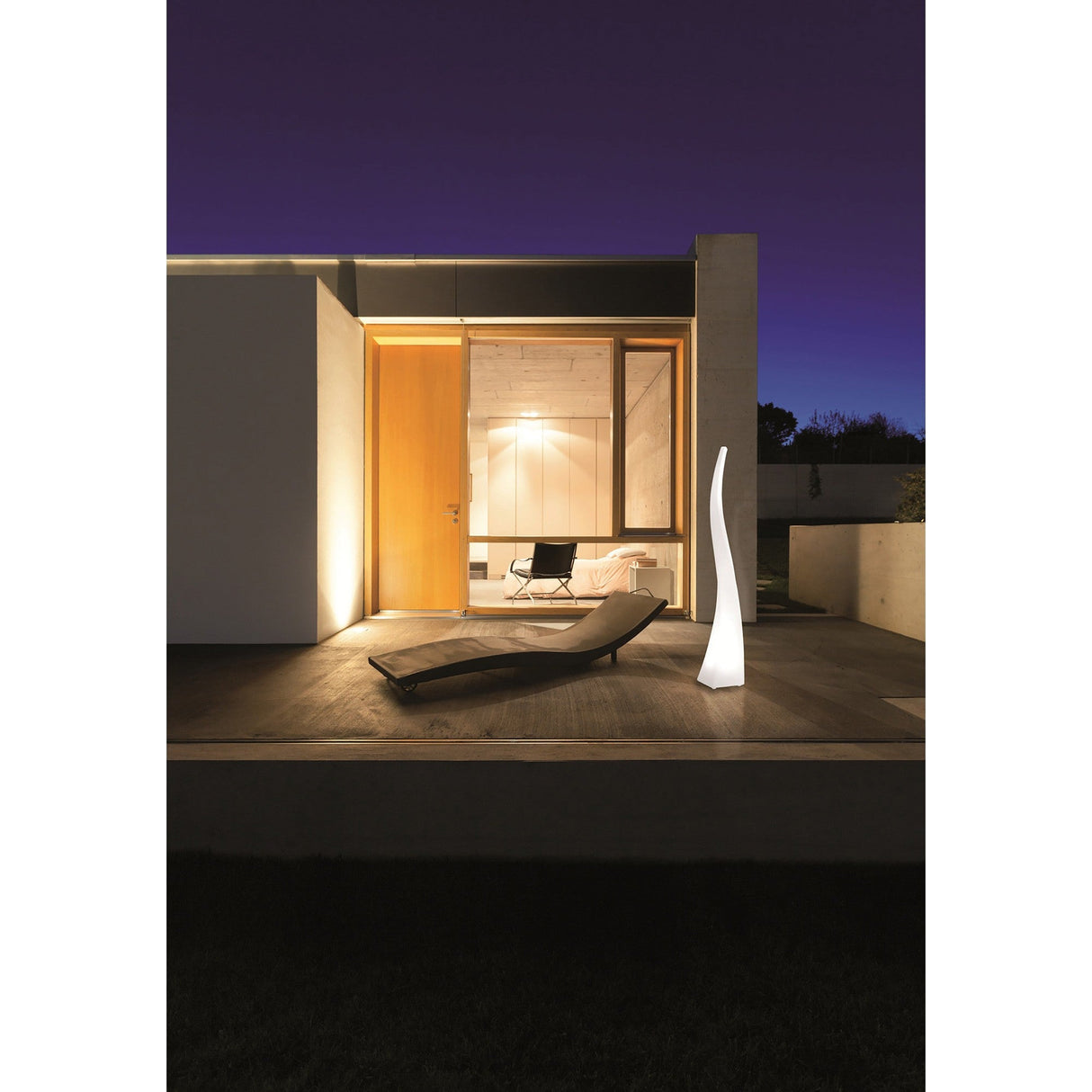 A modern house at night with a large wooden door, glowing window, and a minimalist lounge chair on the deck—enhanced by the Flame 5 Light Outdoor Floor Lamp in Opal White for a stylish touch of weather-resistant lighting.