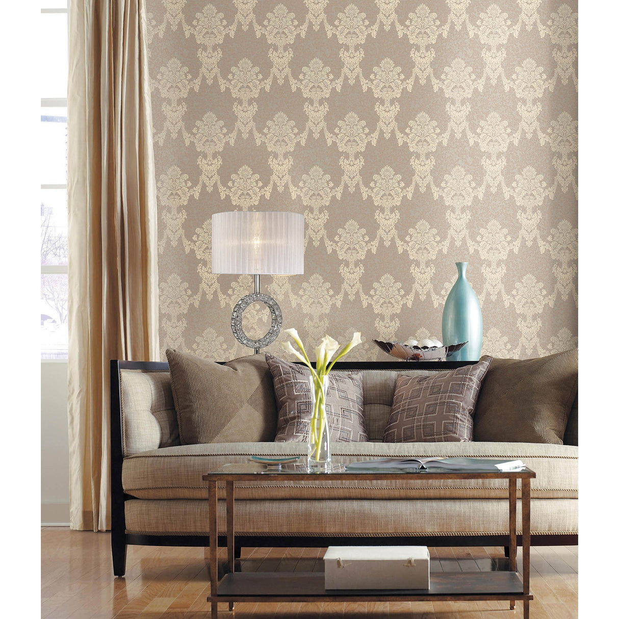 A stylish living room with beige patterned wallpaper, a light sofa, assorted pillows, glass coffee table, a Florence Round 1 Light Crystal Table Lamp - Chrome With White Shade, vase of flowers, and floor-length curtains by the window.