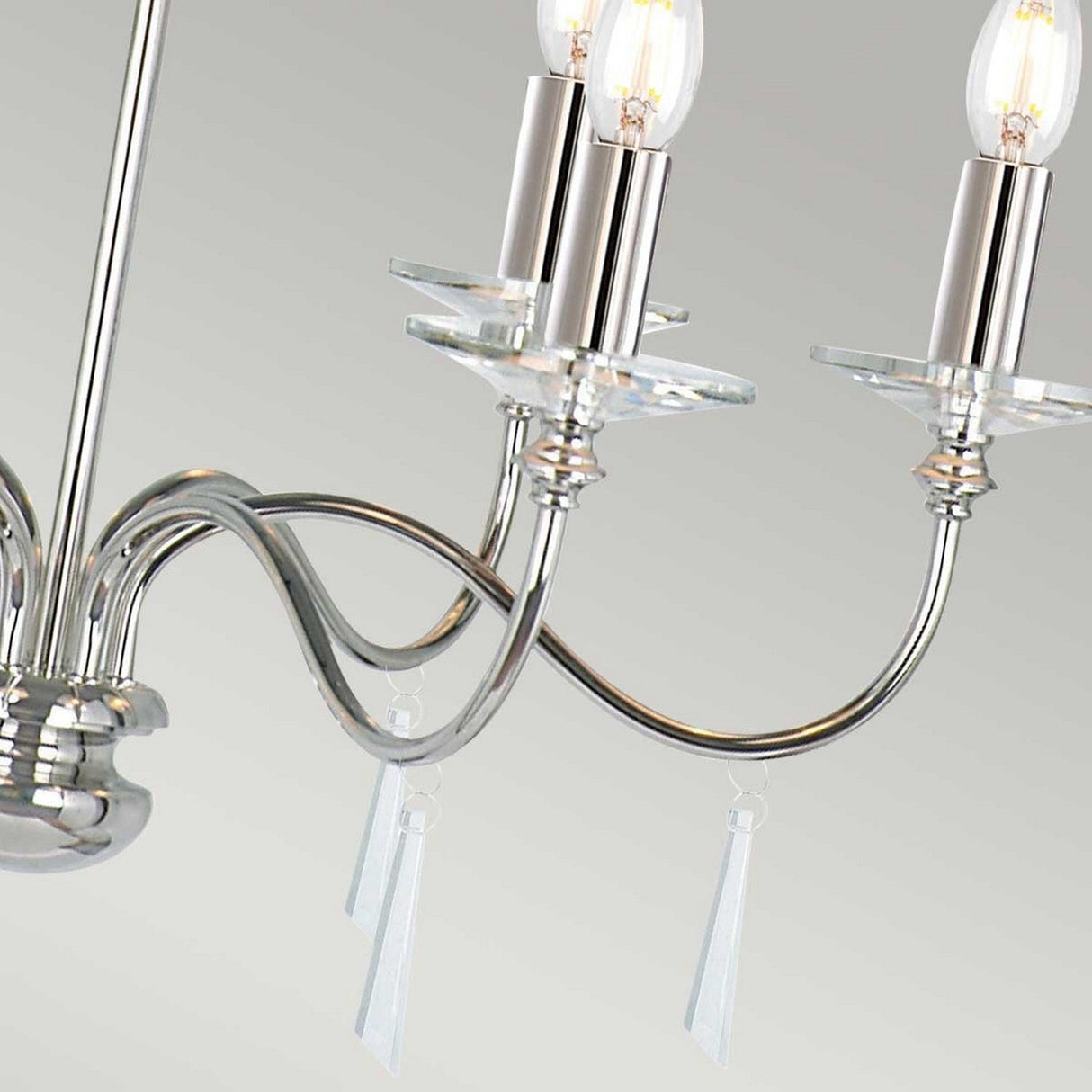 A detailed view of the Finsbury Park 6 Light Chandelier - Polished Nickel highlights its polished finish and graceful, curved arms. Each arm holds two candle-style light bulbs, with hand-cut glass pendants enhancing its sophistication against the simple, light-colored wall.