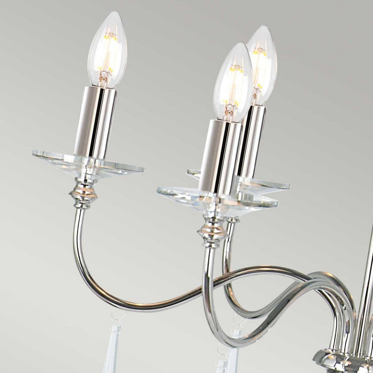 A close-up of the Finsbury Park 6 Light Chandelier in polished nickel showcases its multiple arms and LED bulbs that mimic candle flames, with a sleek reflective finish enhanced by hand-cut glass elements against a soft gray background.