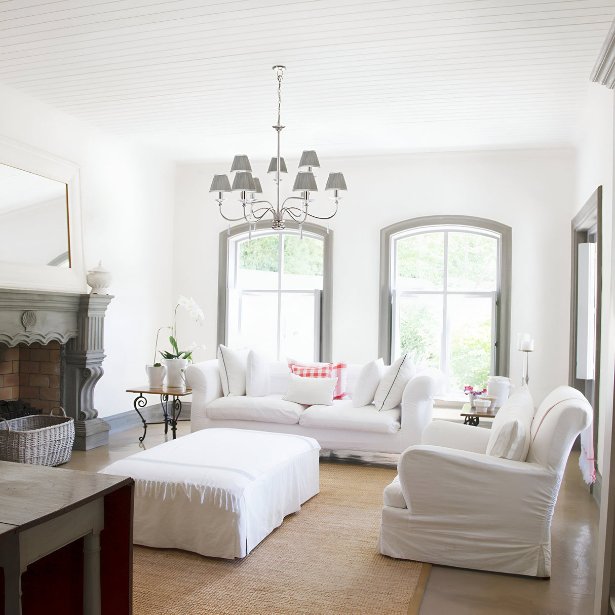 A bright living room with white furniture, including a sofa and armchairs. A soft brown rug covers the floor, while a Finsbury Park 9 Light Chandelier in polished nickel hangs from the ceiling. Large windows allow natural light to stream in, reflecting off hand-cut glass accents by the fireplace on the left.