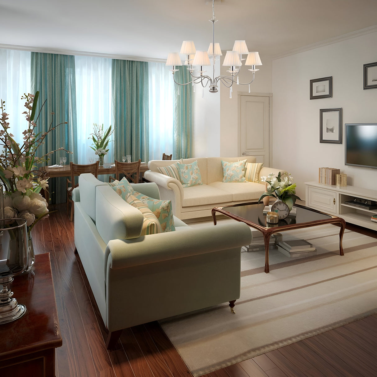 In a welcoming living room, two light gray sofas rest around a wooden coffee table placed on a beige rug. Overhead, the Finsbury Park 9 Light Chandelier in polished nickel casts its glow with intricate hand-cut glass details. Large windows are draped with soft green curtains, and the walls are adorned with floral decorations and framed artwork. A TV is mounted on the right side of the room.