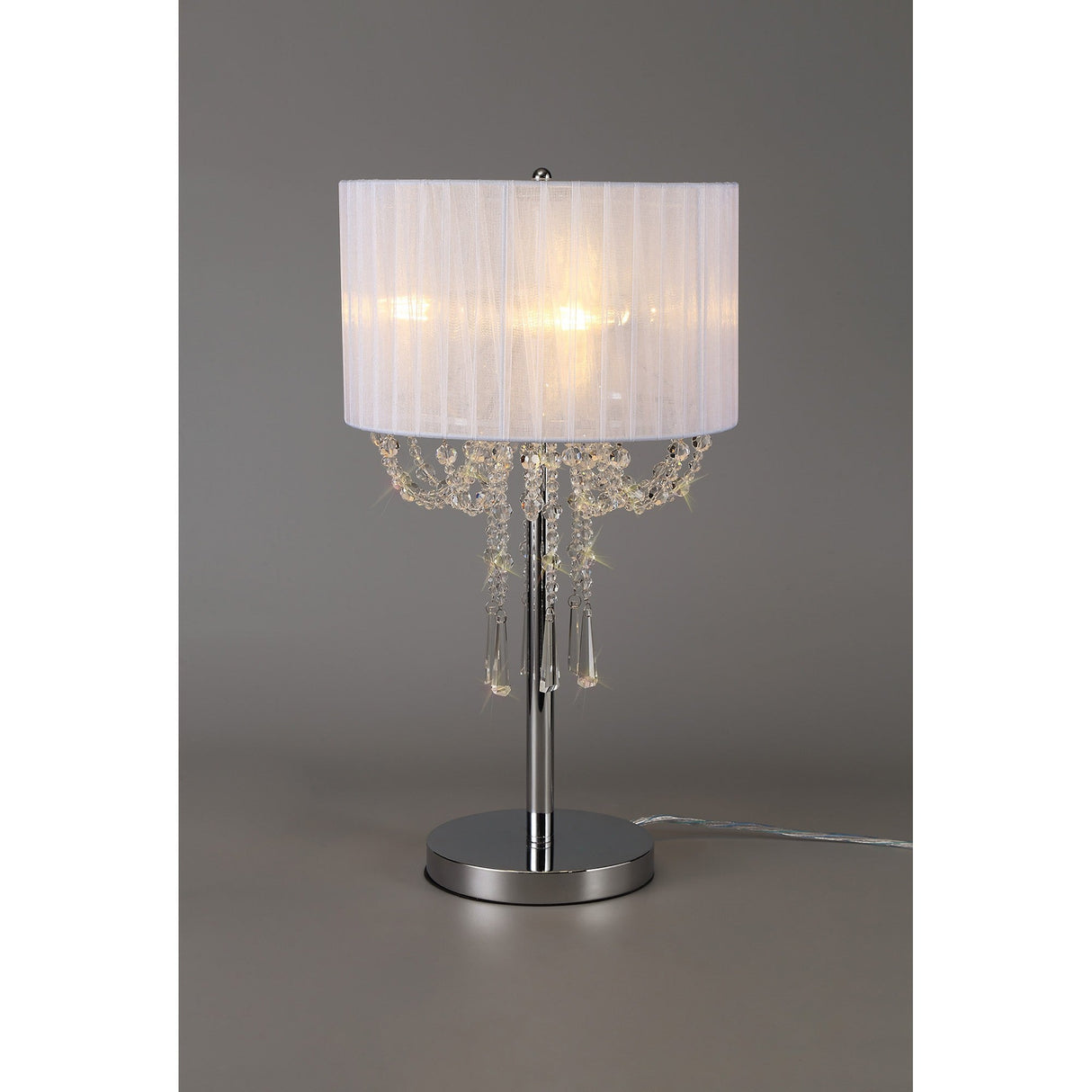 The Freida 3 Light Crystal Table Lamp features a polished chrome frame, a white pleated drum shade, and hanging crystal bead and drop accents. A power cord extends from the base, shown against a plain gray background.