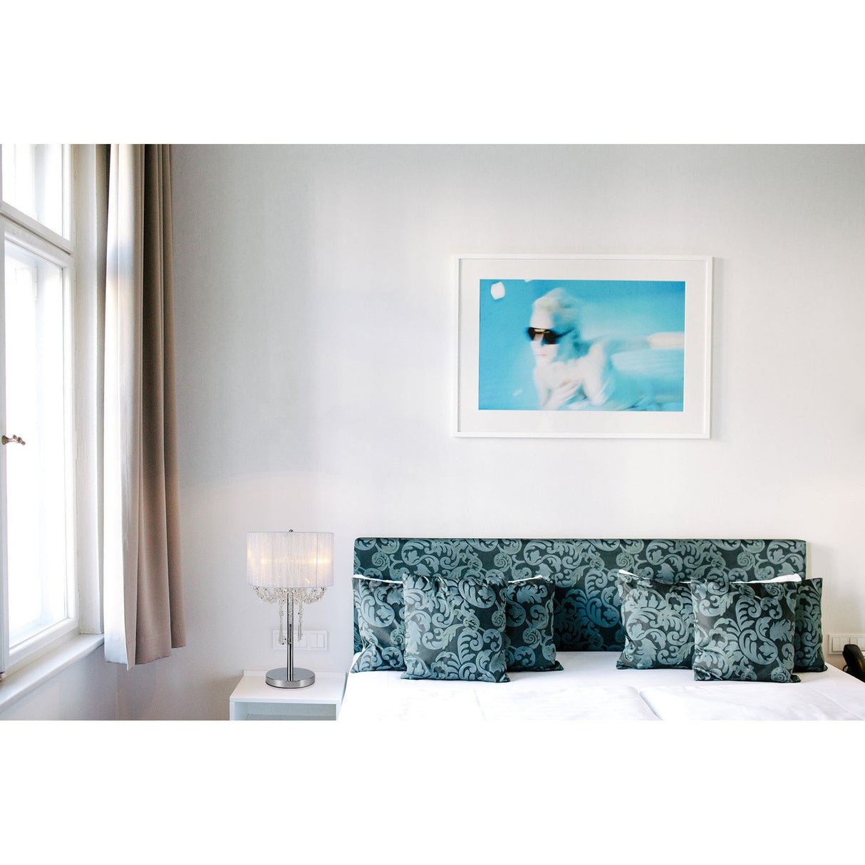 A modern bedroom with a patterned blue headboard, matching pillows, and a bedside table showcasing the Freida 3 Light Crystal Table Lamp - Chrome With White Shade. A large window with curtains and blue-toned artwork complete the look.