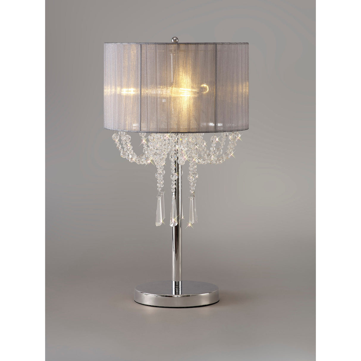 The Freida 3 Light Crystal Table Lamp features a polished chrome finish, grey organza drum shade, and chandelier-like hanging crystal accents, creating a warm glow through its elegant design.