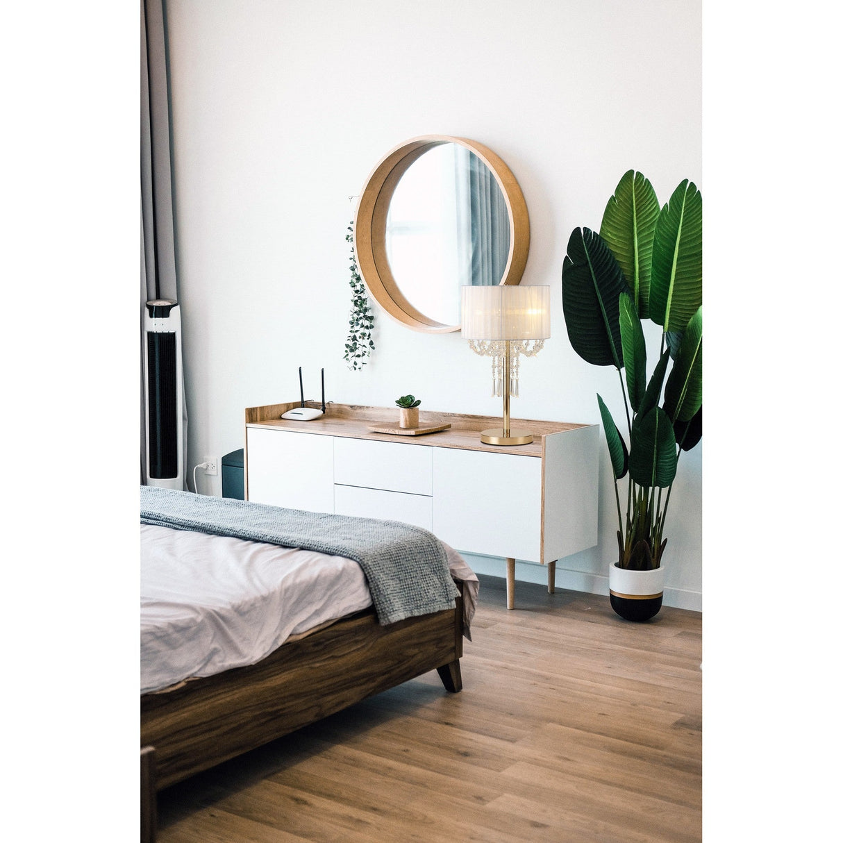 A modern bedroom with a wooden bed, white dresser, round mirror, tall potted plant, and the Freida 3 Light Crystal Table Lamp in gold with a cream shade, set on light wood floors against a white wall.