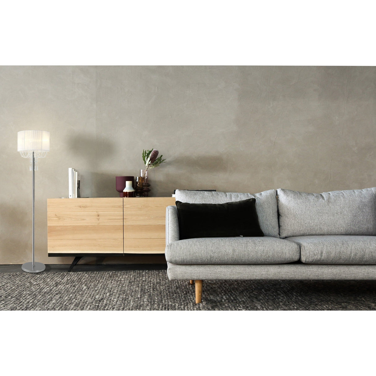 A modern living room featuring a light gray sofa, a wooden sideboard with decor, the Freida 3 Light Crystal Floor Lamp in chrome with a white shade, and a textured gray rug against neutral-toned walls.