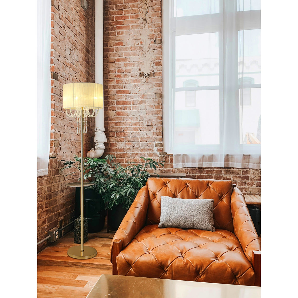 A cozy room with an orange-brown tufted leather armchair, gray pillow, green plants, exposed brick walls, large windows with sheer white curtains, and the Freida 3 Light Crystal Floor Lamp - Gold With Cream Shade illuminating the space.