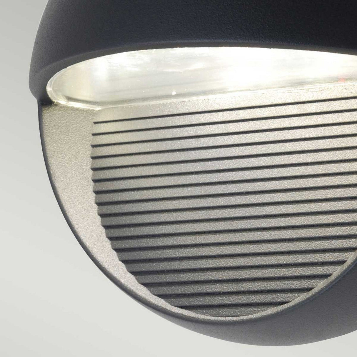 Close-up of the Freyr Outdoor Round Wall Light in graphite, showcasing its modern, circular design with a striped grill on the bottom half. The top half emits bright white light, offering an elegant contemporary lighting solution against a neutral background.