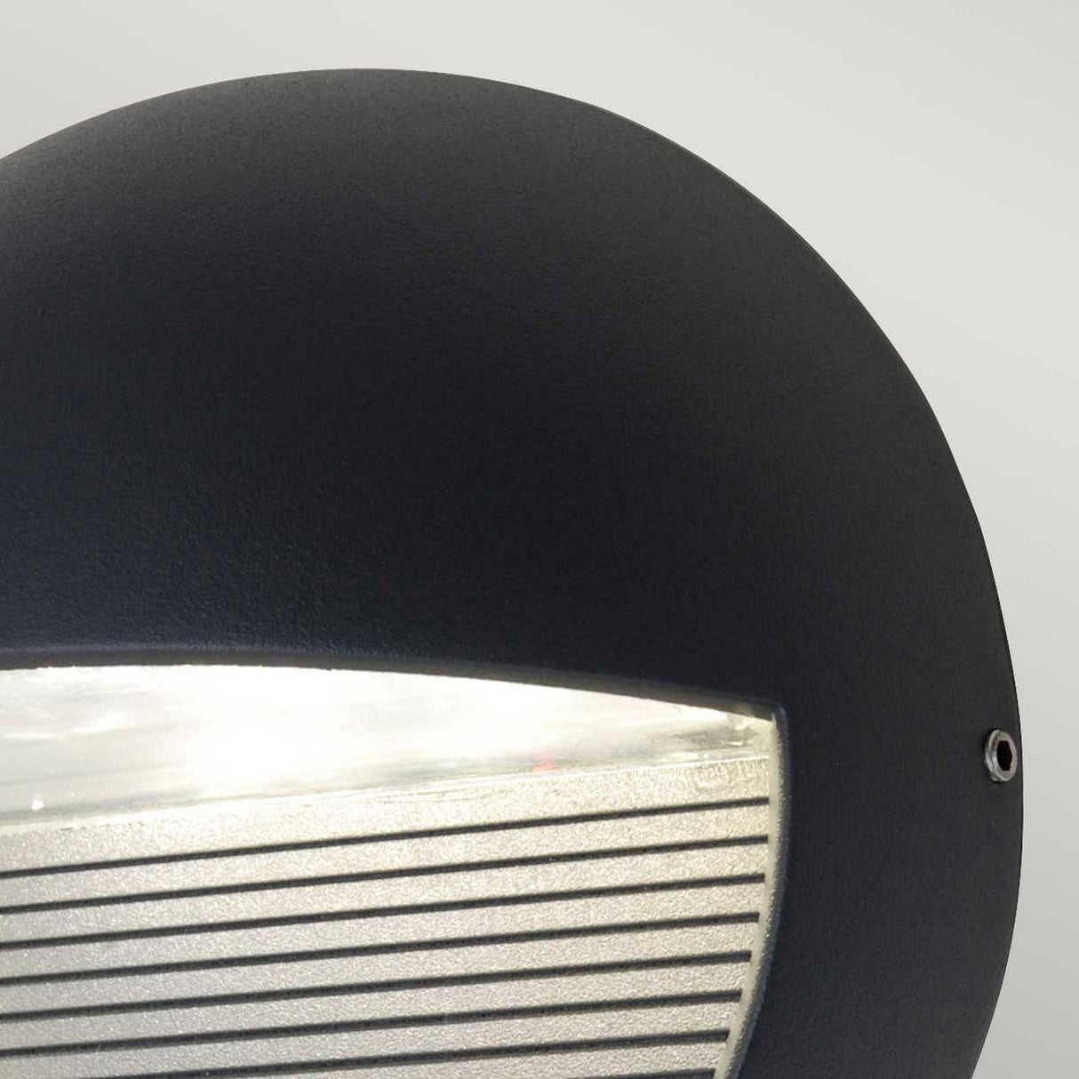 The Freyr Outdoor Round Wall Light in graphite offers a modern lighting solution with its sleek, curved design. Its LED lamp housing emits light from the bottom section, highlighting a lined pattern against a soft gray background.