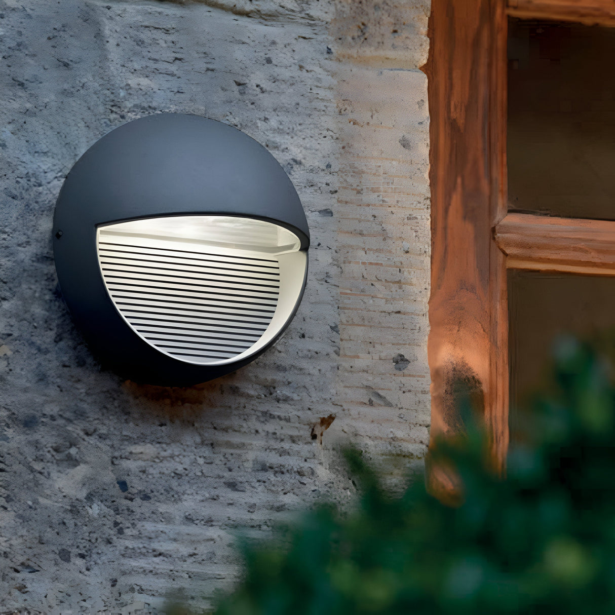 The Freyr Outdoor Round Wall Light in a stylish graphite finish features a half-sphere design on a stone wall, positioned adjacent to a wooden window frame. The LED lamp emits a warm downward glow, with greenery subtly appearing at the lower right corner of the image.