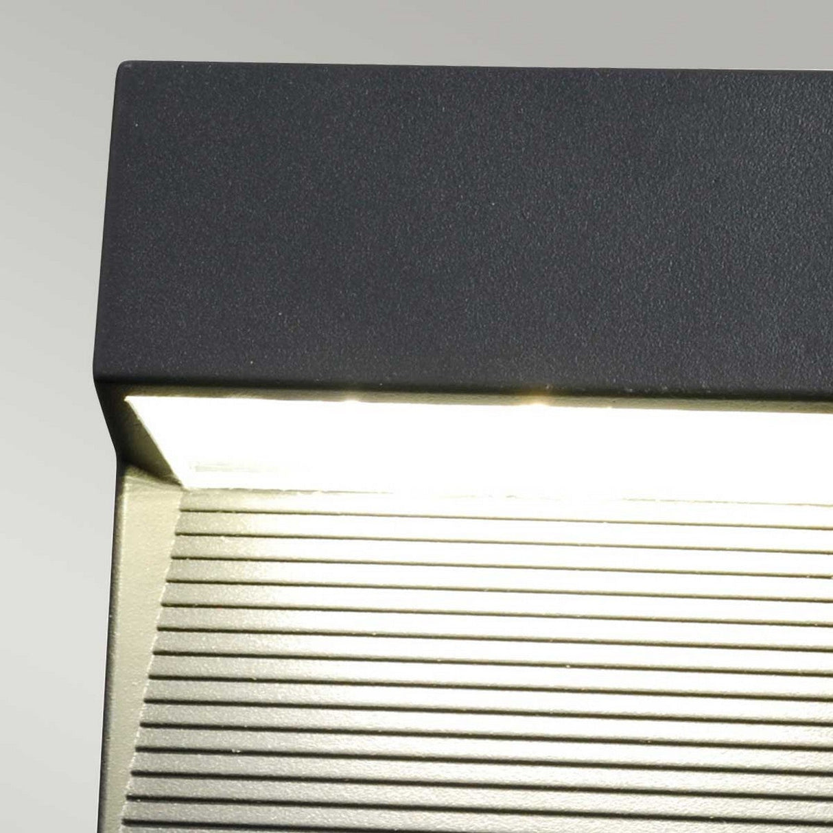 Close-up of the Freyr Outdoor Square Wall Light in graphite, crafted from durable die-cast aluminum. The top part is a dark rectangular casing, while the lower section features horizontal grooves illuminated by bright white light against a plain light gray background.