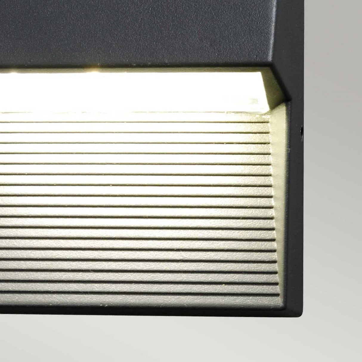 A detailed view of the Freyr Outdoor Square Wall Light - Graphite highlights its sturdy die-cast aluminum frame and horizontal slats. Its modern design casts a warm glow against a simple backdrop, seamlessly merging contemporary aesthetics with practical function.