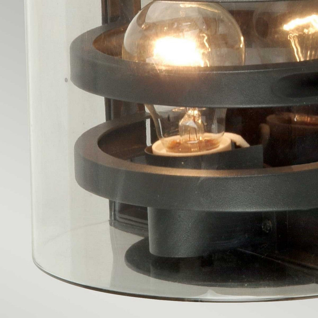 Close-up of the Frida Outdoor Wall Light - Graphite, showcasing a modern cylindrical design with visible incandescent bulbs. The fixture is crafted from sleek die-cast aluminium and covered with transparent glass, highlighting the glowing bulbs inside and complemented by its graphite metal accents.