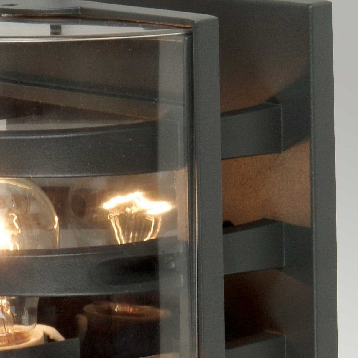 Close-up of the Frida Outdoor Wall Light in graphite, featuring a glass cover that showcases an exposed filament bulb. The fixture's angular design in die-cast aluminium and reflective surfaces are prominently highlighted, with a visible glow that exemplifies modern lighting design.