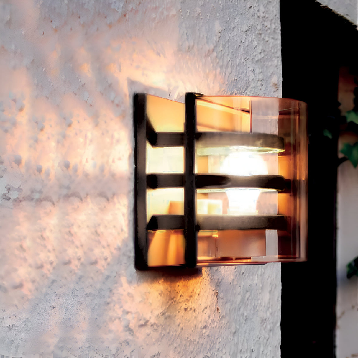 The Frida Outdoor Wall Light in graphite, crafted from die-cast aluminum with a unique grid design, emits a warm glow against a textured white wall, casting subtle shadows.