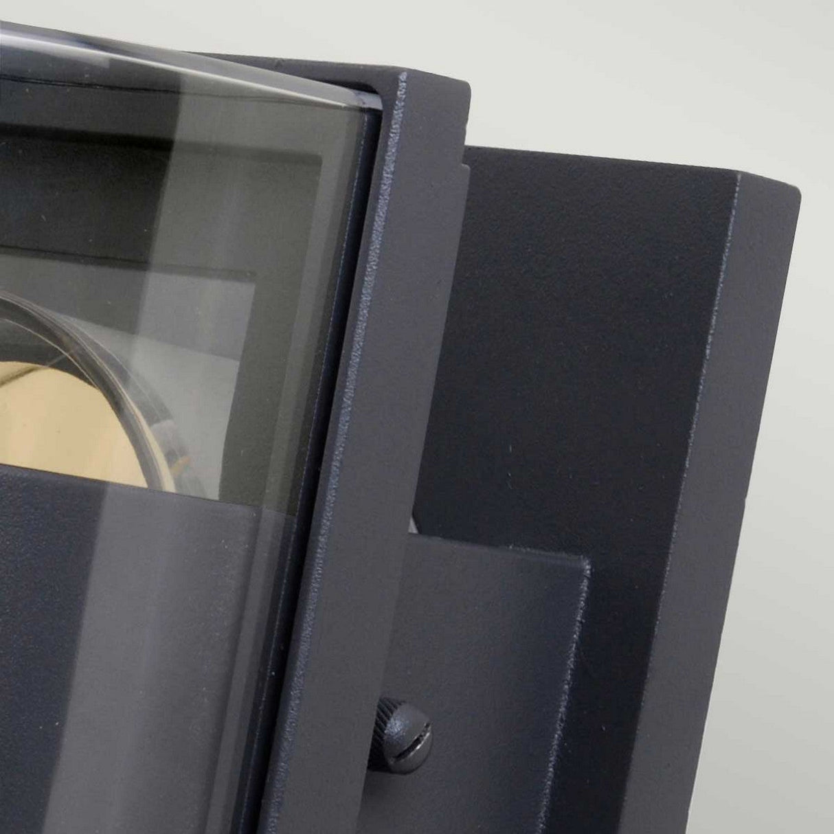 A close-up of the Frida Outdoor Small Wall Light in graphite reveals its black, angular design with a clear glass front, showcasing a metallic interior that is likely made from die-cast aluminium. Set against a neutral gray background, this sleek fixture suggests durability with its IP54 rating.