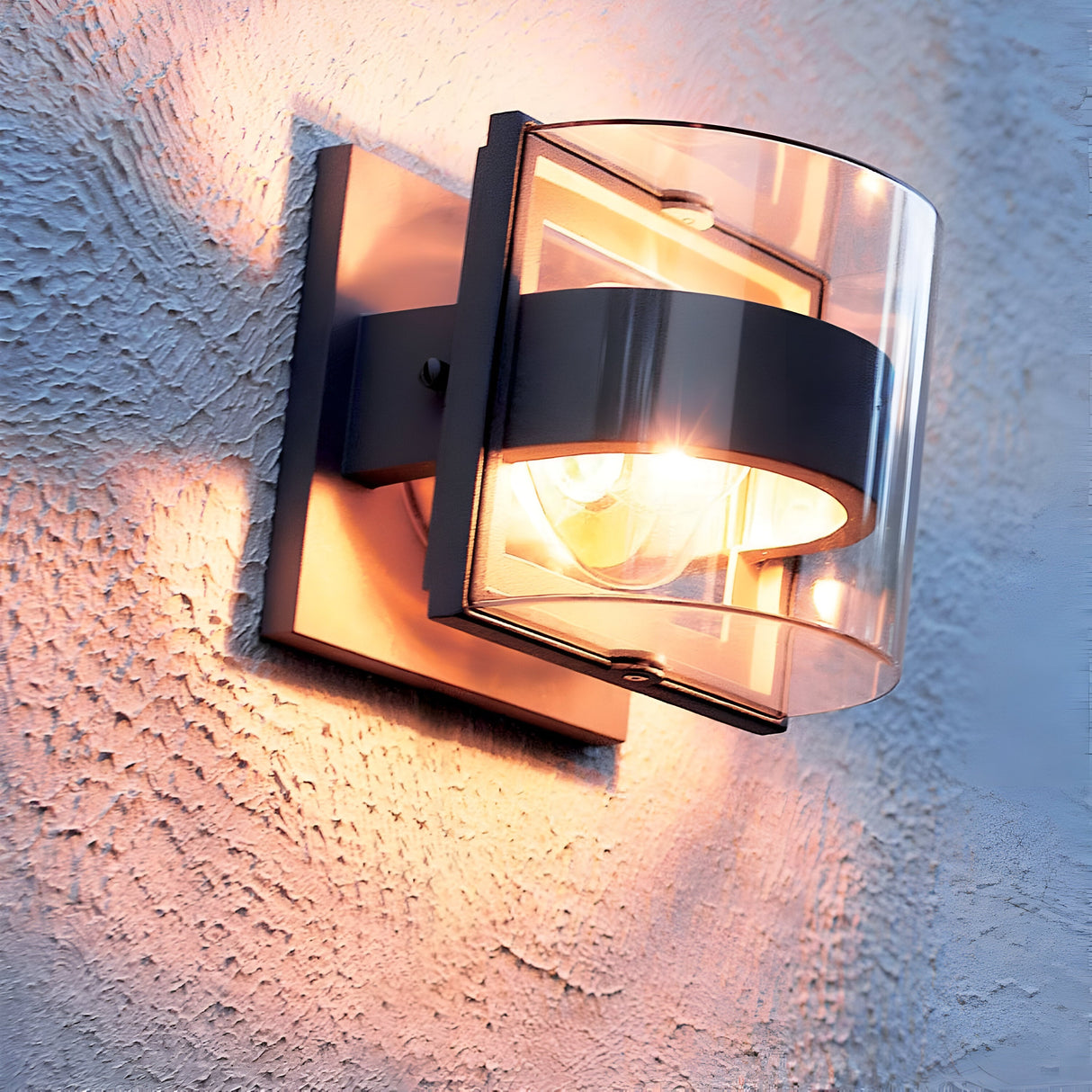 The Frida Outdoor Small Wall Light - Graphite, with its clear cylindrical glass shade and die-cast aluminum construction, is mounted on a textured wall. Boasting an IP54 rating, it creates a warm and inviting atmosphere while casting soft shadows across the surface.