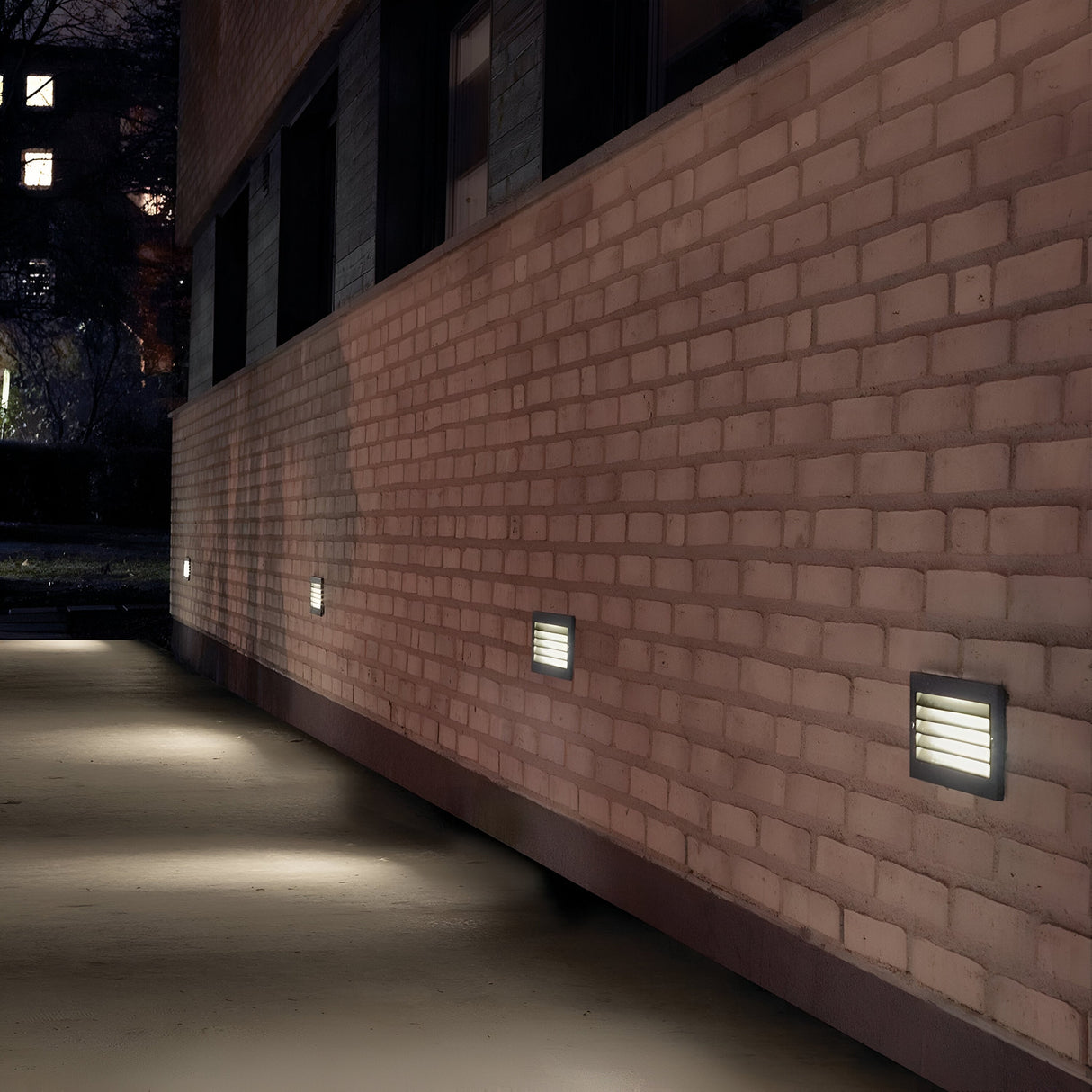 A nighttime scene of a brick building features several Grimstad LED Recessed Wall Lights in graphite embedded along a pathway. The energy-efficient lighting illuminates the path, while trees and other buildings create a picturesque backdrop.