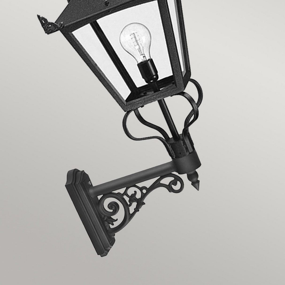 The Grampian 1 Light Outdoor Wall Lantern in black showcases a decorative bracket and a visible bulb inside, crafted from cast aluminum. This Victorian-inspired design is striking against a plain gray background, emphasizing its intricate details and classic style.