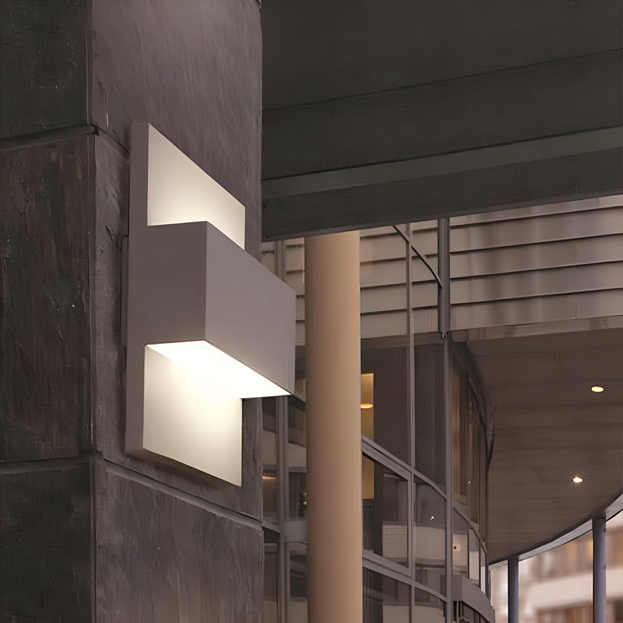 The Geneve Outdoor 1 Light Wall Lantern in Silver softly illuminates the dark stone wall with its modern, rectangular design. Made from die-cast aluminum, it perfectly complements large glass windows and columns, evoking an urban architectural aesthetic.