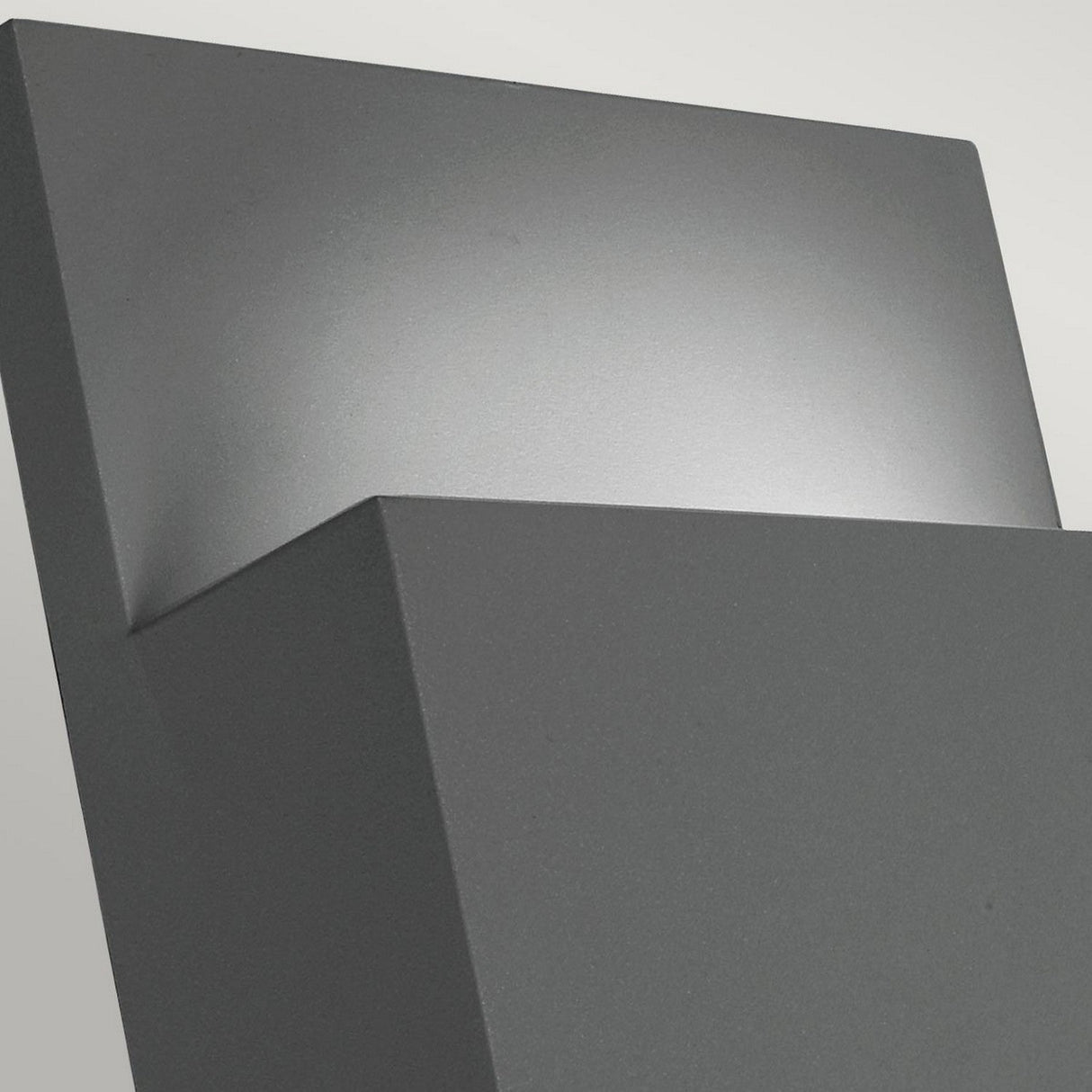 A close-up of the Geneve Outdoor 1 Light Wall Lantern reveals its modern angular design, housed in a sleek graphite finish casing that casts a soft glow against the wall.