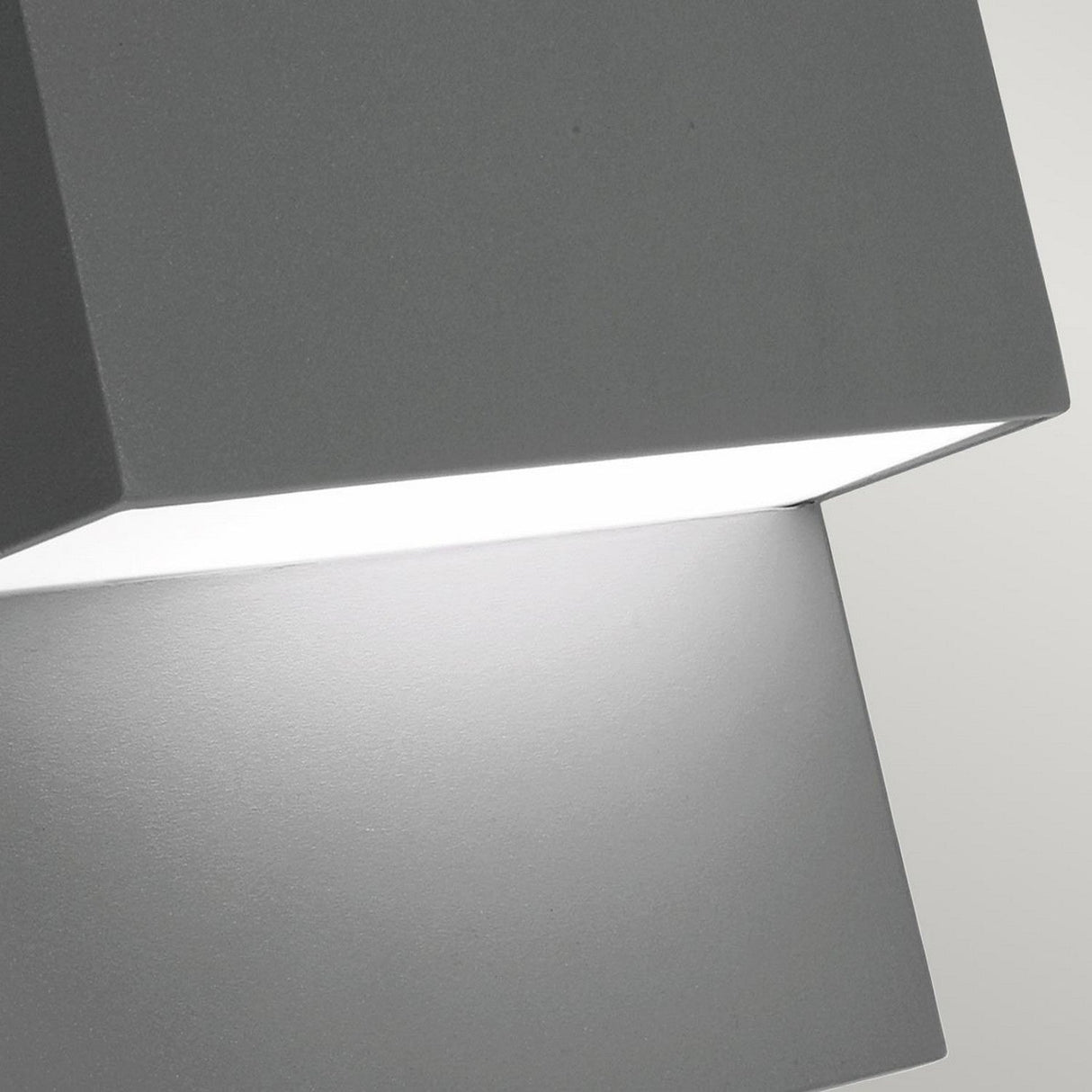 Close-up view of the Geneve Outdoor 1 Light Wall Lantern featuring a graphite finish and angular design. The lantern emits a bright white light, highlighting the contrast between the illuminated portion and the shadowed area.