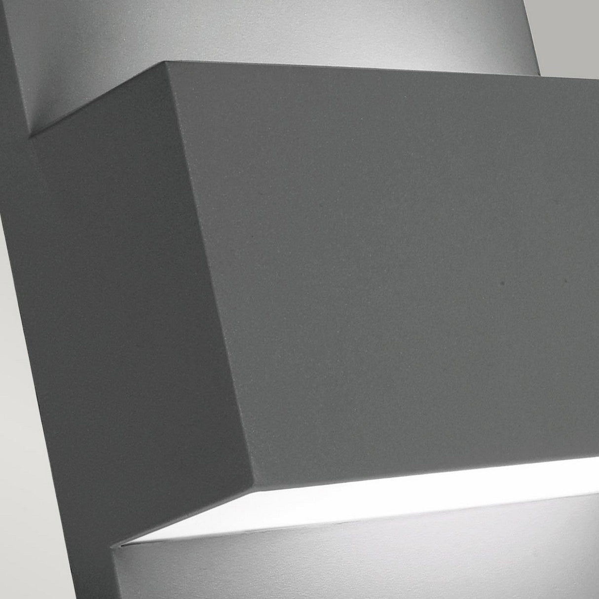 Close-up of the Geneve Outdoor 1 Light Wall Lantern in graphite, featuring a modern geometric rectangular design. This gray wall-mounted light fixture emits a soft white light from its bottom edge, presenting a sleek and minimalist appearance. Ideal for anyone looking for contemporary outdoor wall lanterns or modern lighting solutions.