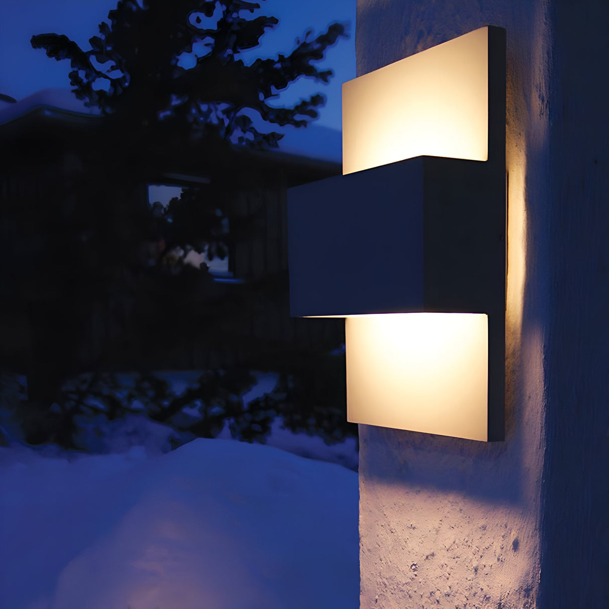 The Geneve Outdoor 1 Light Wall Lantern in a sleek graphite finish illuminates the evening, casting a warm glow on the snow-covered area as a tree silhouette stands in the background against the twilight sky.
