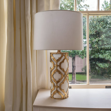 The Arabella 1 Light Table Lamp - Gold With Ivory Shade is gracefully placed on a white table by the window. Its ivory shade harmonizes with the cream curtains, while the lush green trees outside contribute to the tranquil ambiance.