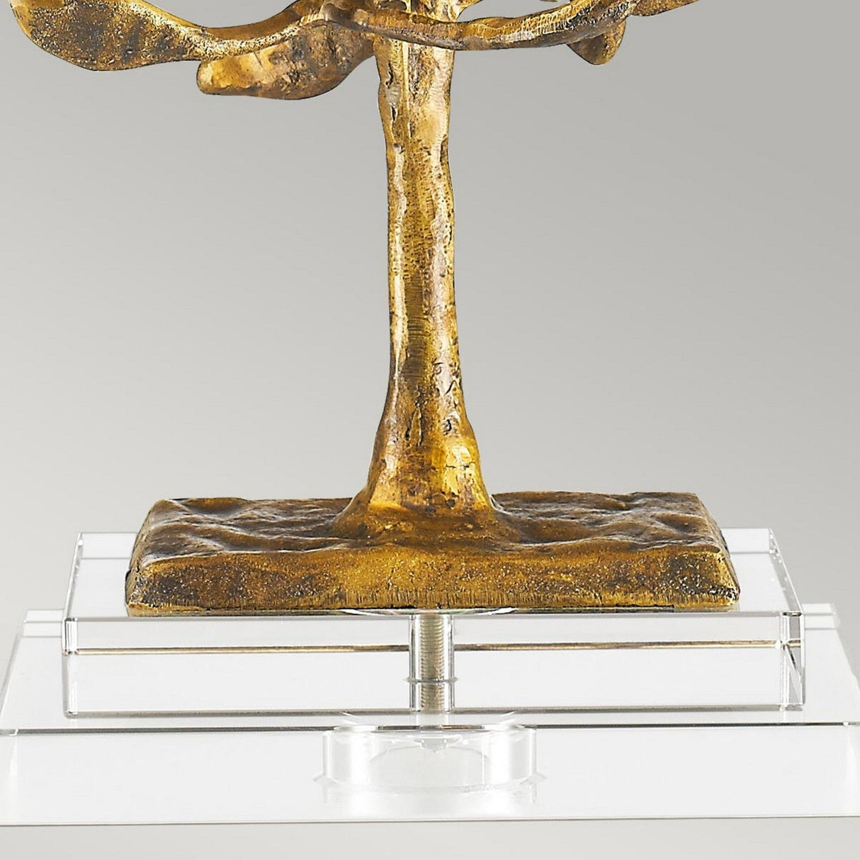 Close-up of the Audubon Park 1 Light Table Lamp - Gold, showcasing a gold cast metal sculpture with a tree motif and textured trunk, mounted on a transparent K9 glass base. The background is plain light gray.