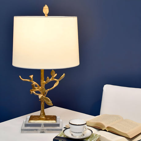An Audubon Park 1 Light Table Lamp in gold with a white shade characterized by a subtle tree motif rests on a white table against a dark blue wall. Beside it, there's a stack of books and an open book next to an immaculate white teacup and saucer.