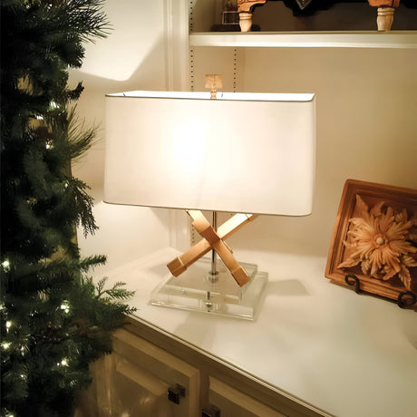 A contemporary Jackson Square 1 Light Table Lamp in gold, with a cream linen shade and intersecting wooden rods, is elegantly positioned on a clear base. It adorns a white cabinet placed beside a glowing Christmas tree and an artfully framed decorative piece on the shelf.